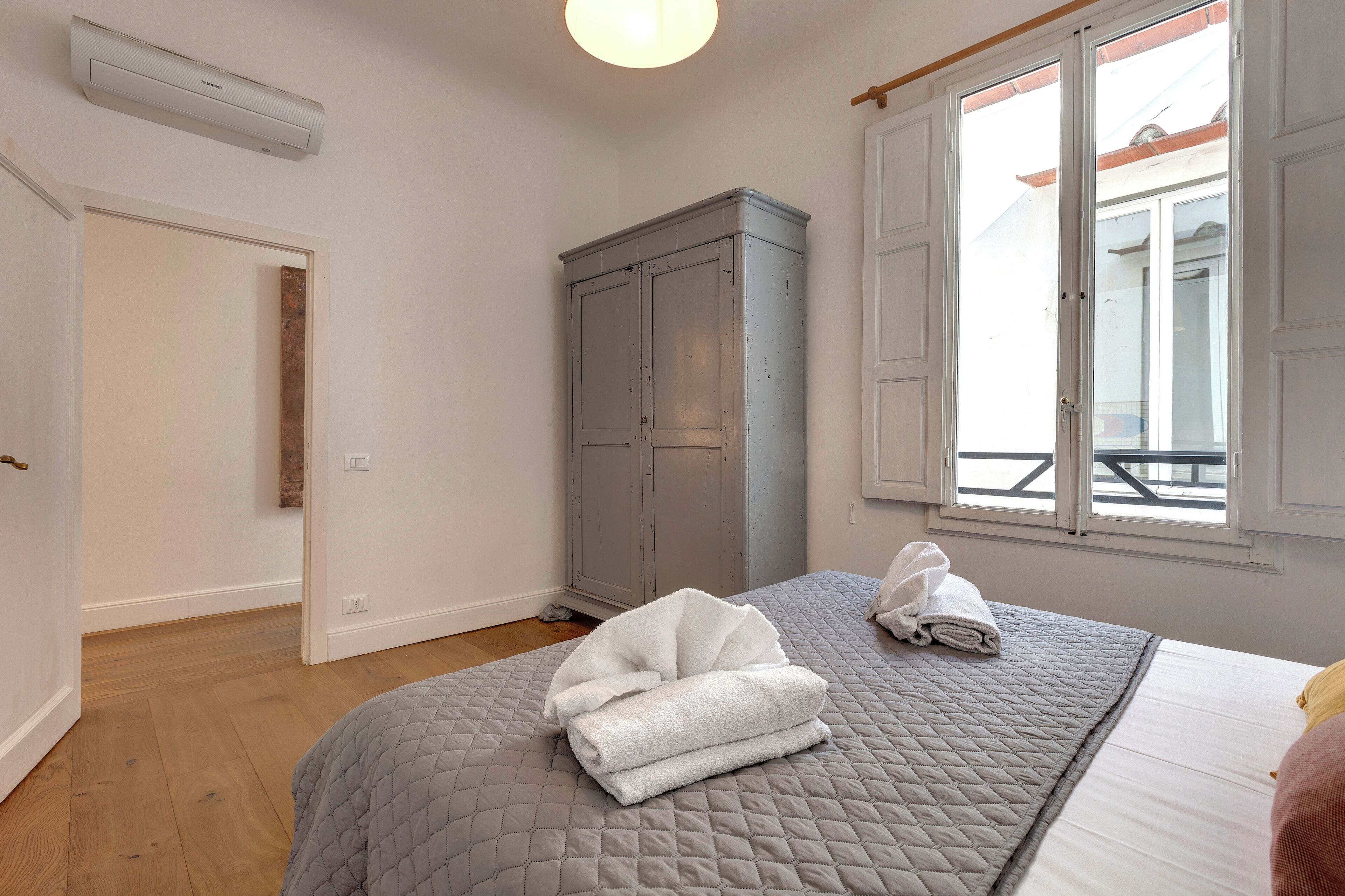 Ein Schlafzimmer mit einer grauen gesteppten Bettdecke, einem grauen Kleiderschrank und einem Fenster mit Blick auf einen Balkon.