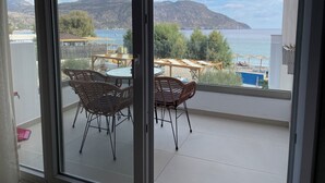 A balcony with wicker chairs and a table, overlooking a beach and mountains. - Tamaris Suites (Karpathos)