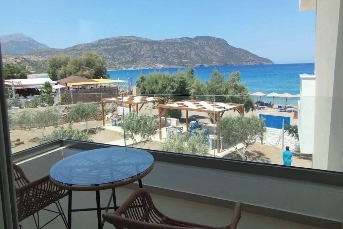 A balcony with a glass table and wicker chairs, overlooking a pool area with umbrellas and lounge chairs, and a view of the sea and mountains.