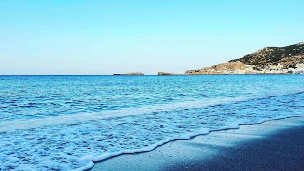 A beach with waves gently lapping onto the shore, a clear sky, and a rocky coastline in the background. - Tamaris Suites (Karpathos)