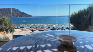 A beach view from a room with a glass partition, featuring a clear view of the sea, beach umbrellas, and lounge chairs. - Tamaris Suites (Karpathos)