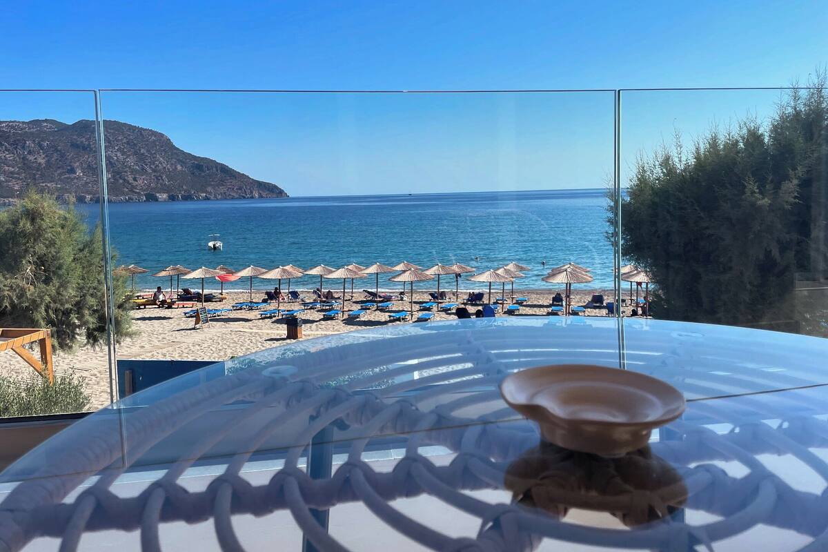 A beach view from a room with a glass partition, featuring a clear view of the sea, beach umbrellas, and lounge chairs.