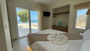 A hotel room with a bed, a TV, a window with a view of the beach, and a door leading to a balcony. - Tamaris Suites (Karpathos)