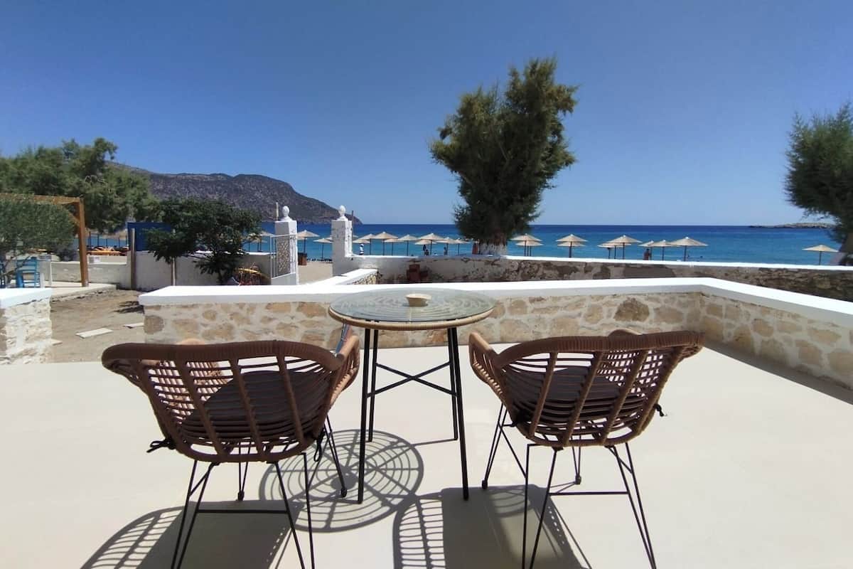 A terrace with two wicker chairs and a table, overlooking a beach with umbrellas and a mountain in the background.