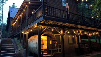 A wooden cabin with a circular interior structure, a balcony with string lights, and a staircase leading to the entrance.