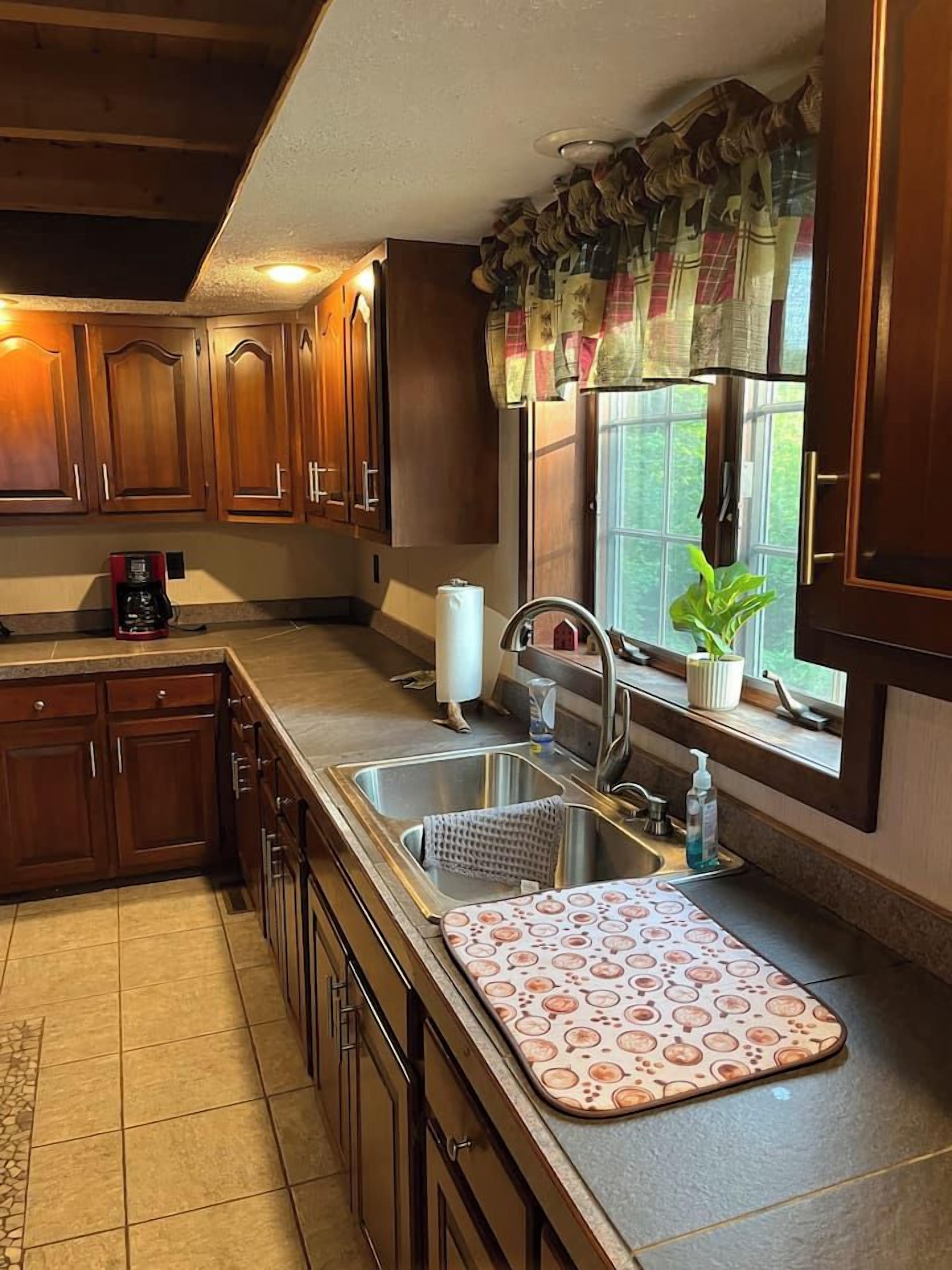 A kitchen with wooden cabinets, a stainless steel sink, a coffee maker, and a window with a curtain.