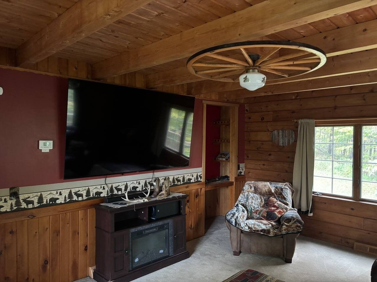 A wooden cabin interior with a large flat-screen TV mounted on the wall, a fireplace, a patterned armchair, and a ceiling fan.