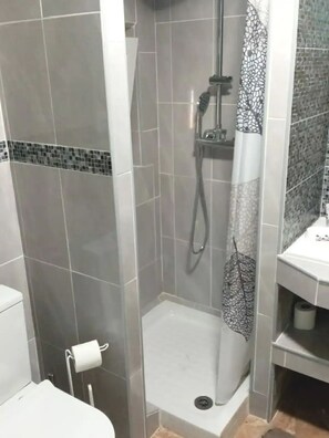 A bathroom with grey tiled walls, a shower with a patterned curtain, and a toilet with a roll of toilet paper. - Guesthouse Milagre Centro Faro (Faro)