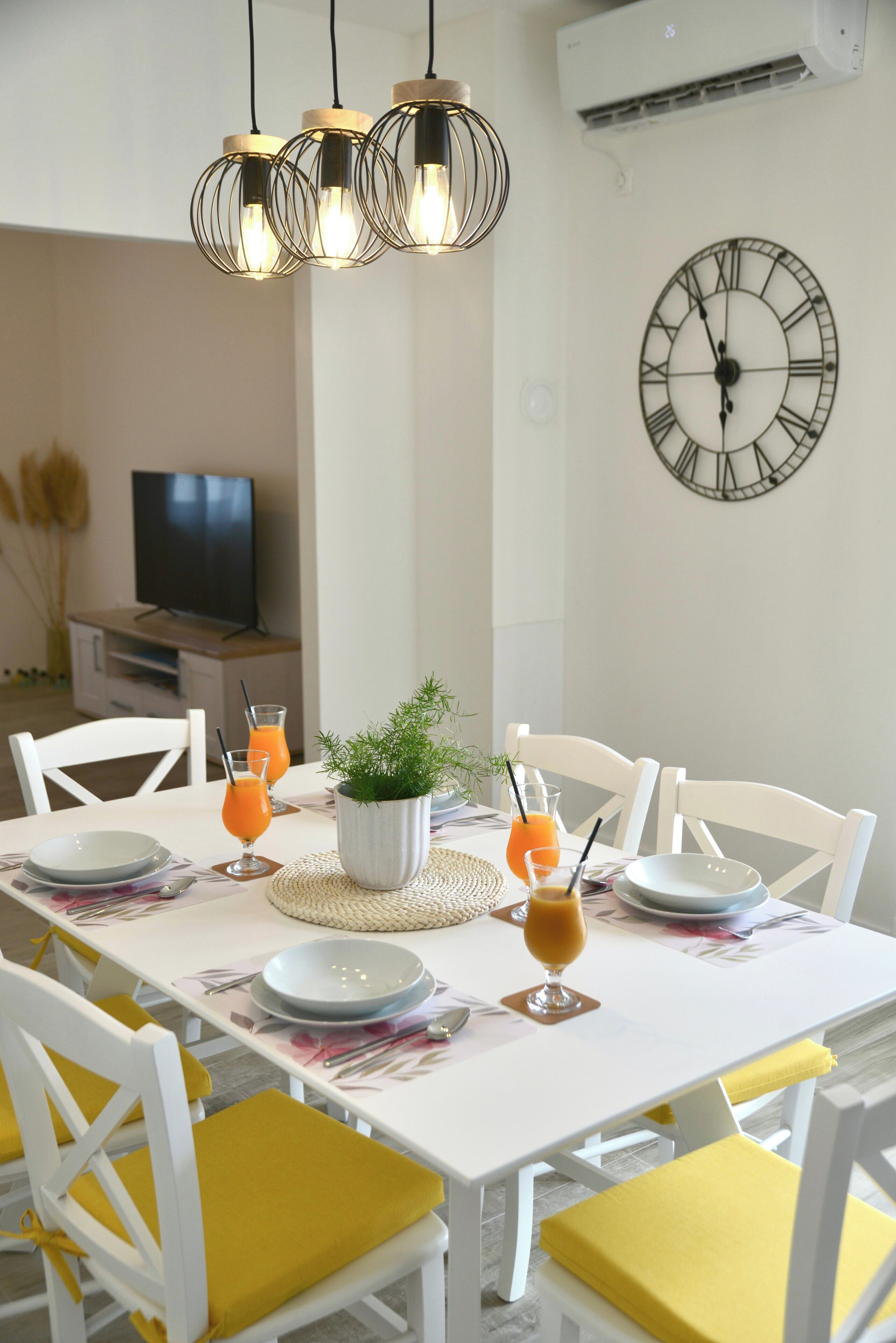 Un coin repas avec une table blanche dressée pour six personnes, des chaises jaunes et une horloge au mur.