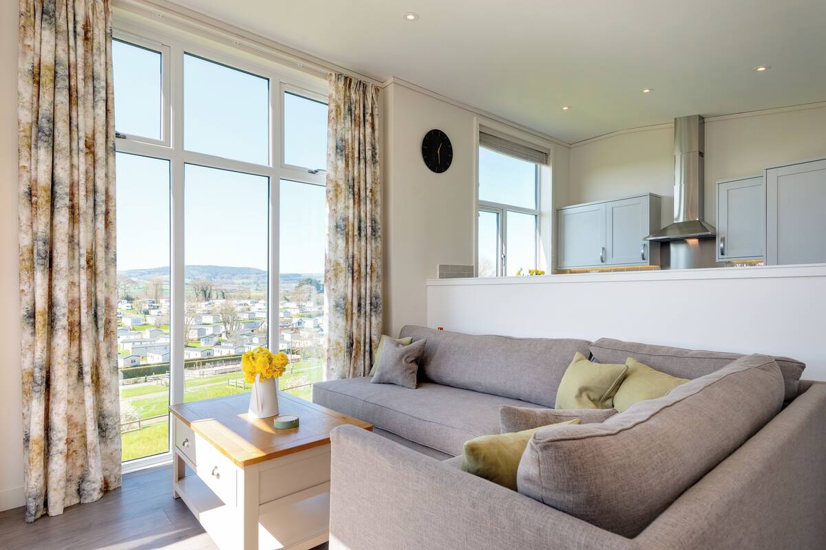 Ein modernes Wohnzimmer mit einem grauen Sofa, einem Holztisch und Blick auf eine Wohngegend durch große Fenster.
