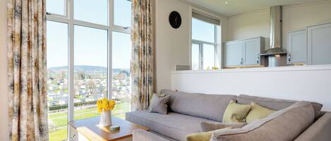 A modern living room with a grey sofa, a wooden side table, and a view of a residential area through large windows.