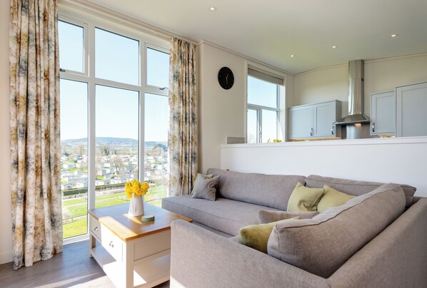 A modern living room with a grey sofa, a wooden side table, and a view of a residential area through large windows.