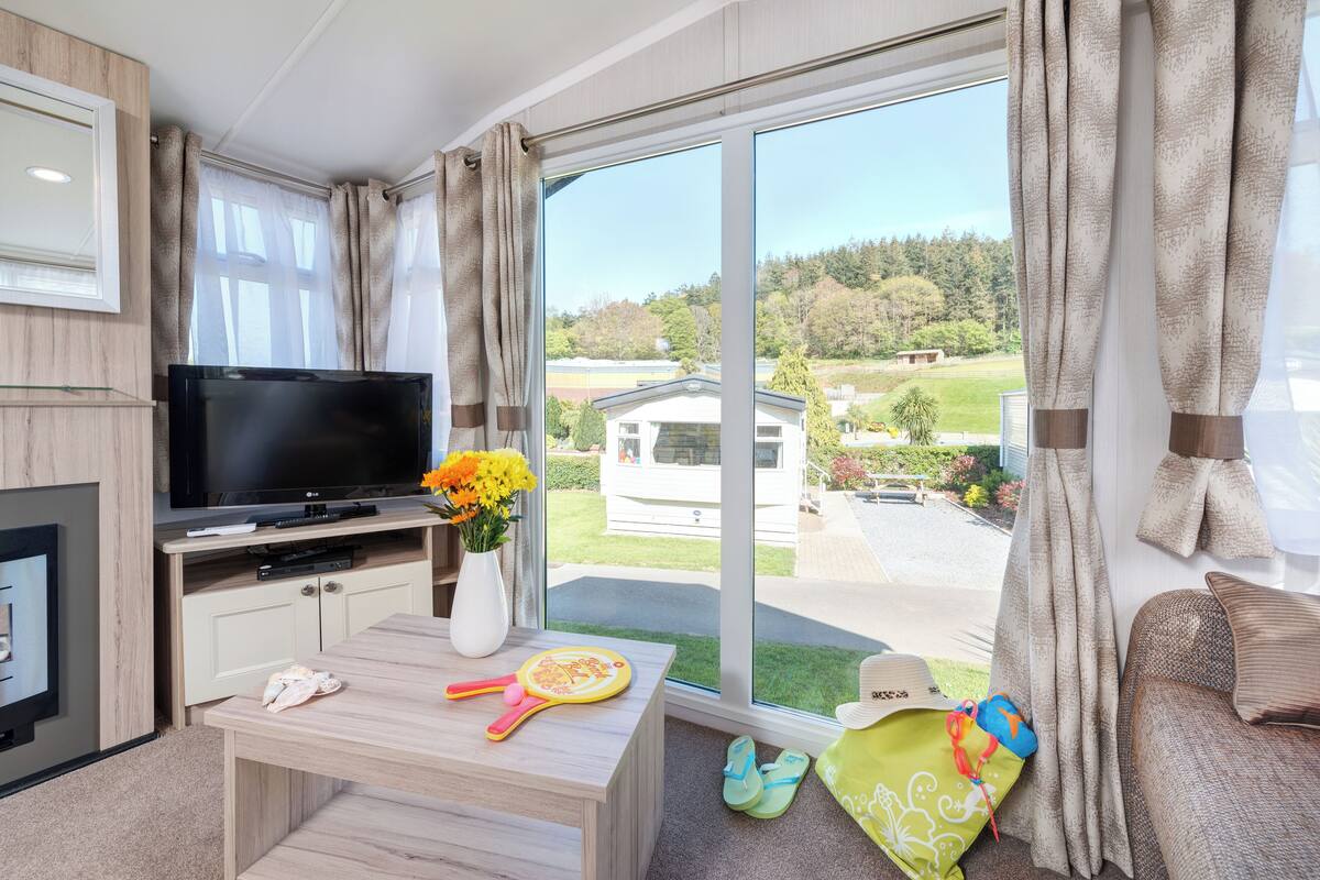 Ein Zimmer mit Fernseher, einem kleinen Tisch mit Blumenvase, einer Couch und einem Fenster mit Vorhängen.