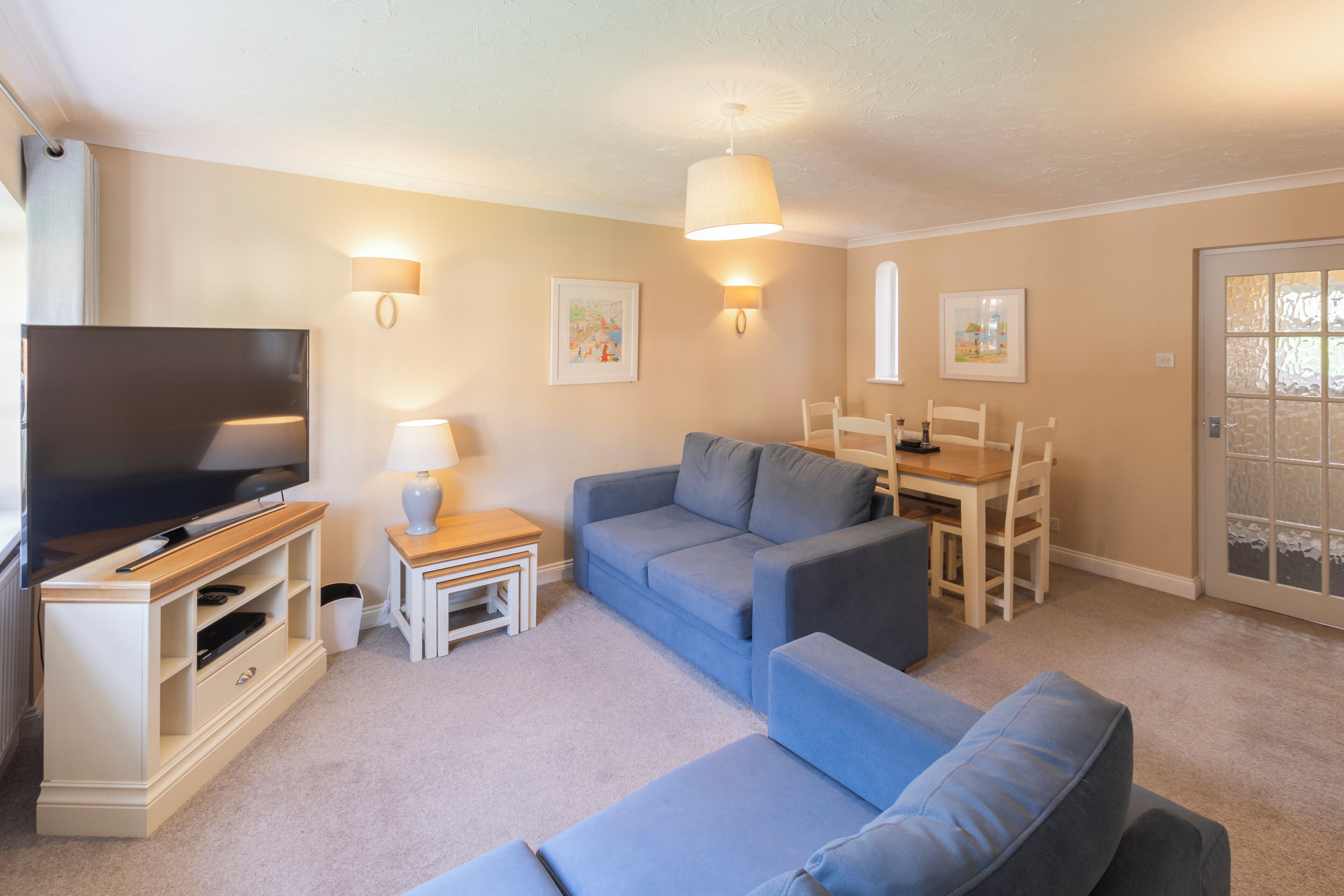 A living room with a flat-screen TV, a blue sofa, a wooden coffee table, and a dining table with chairs.