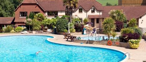 A swimming pool with a person swimming, surrounded by a building with a tiled roof and a garden with various plants.