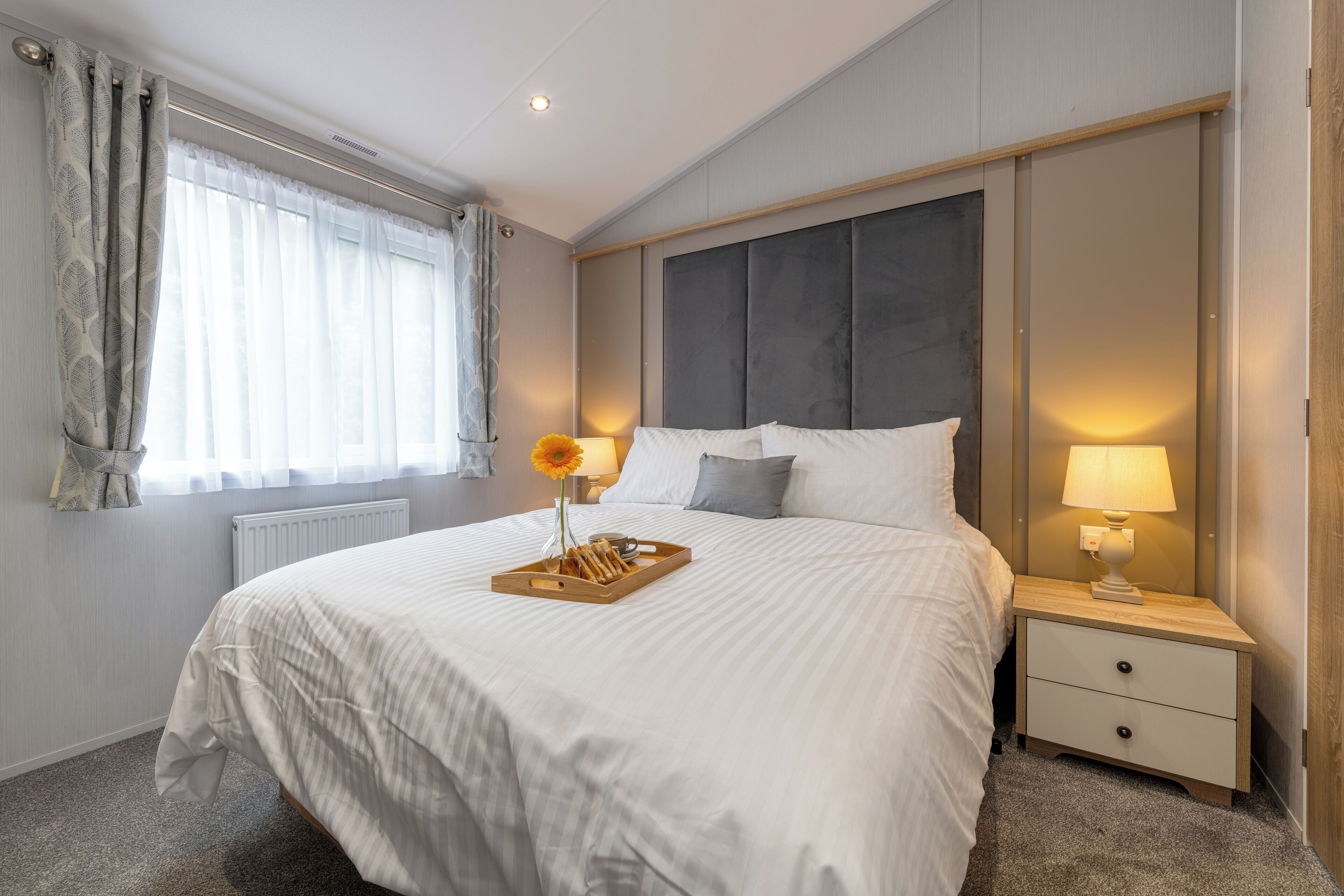A neatly made bed with a tray of bread and a vase of flowers, a bedside lamp, and a nightstand with drawers.