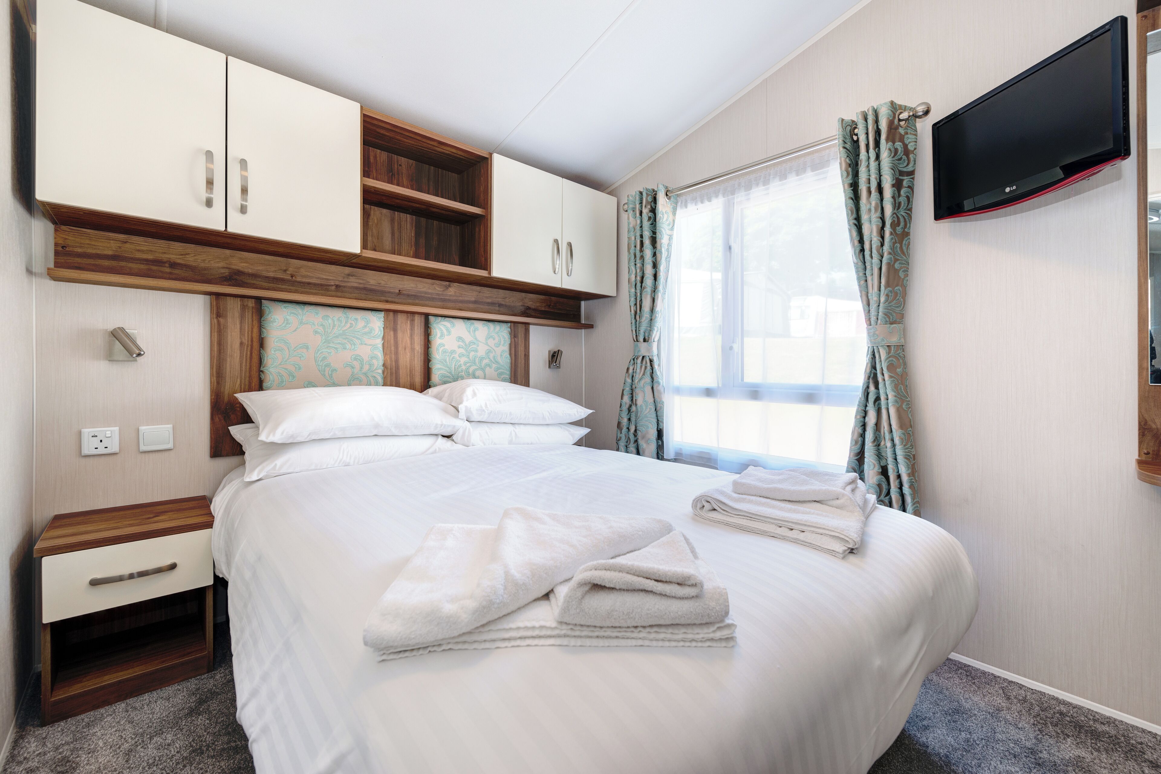 Un lit bien fait, avec du linge de lit et des serviettes blanches, une table de chevet en bois avec un tiroir, une étagère intégrée au-dessus du lit, un téléviseur à écran plat fixé au mur et une fenêtre avec des rideaux.