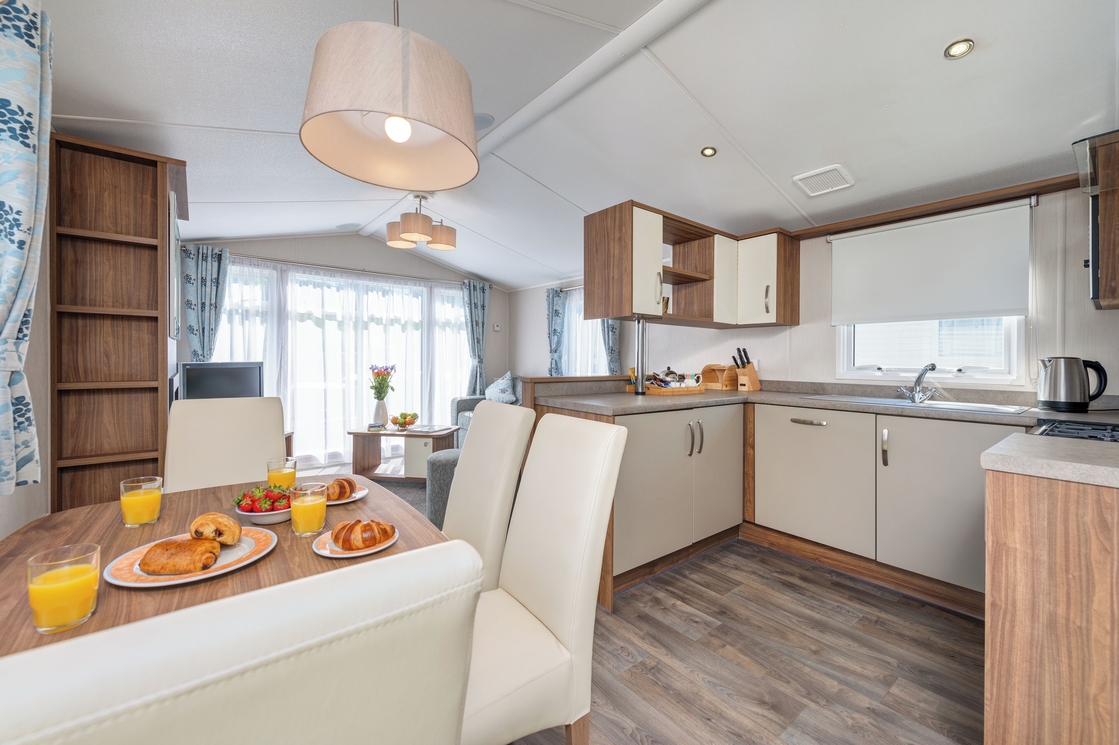 A modern kitchen with wooden cabinets, a dining table with breakfast items, and a large window with curtains.