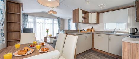 Une cuisine moderne avec des meubles en bois, une table à manger où sont disposés des aliments pour le petit-déjeuner, et une grande fenêtre avec des rideaux.