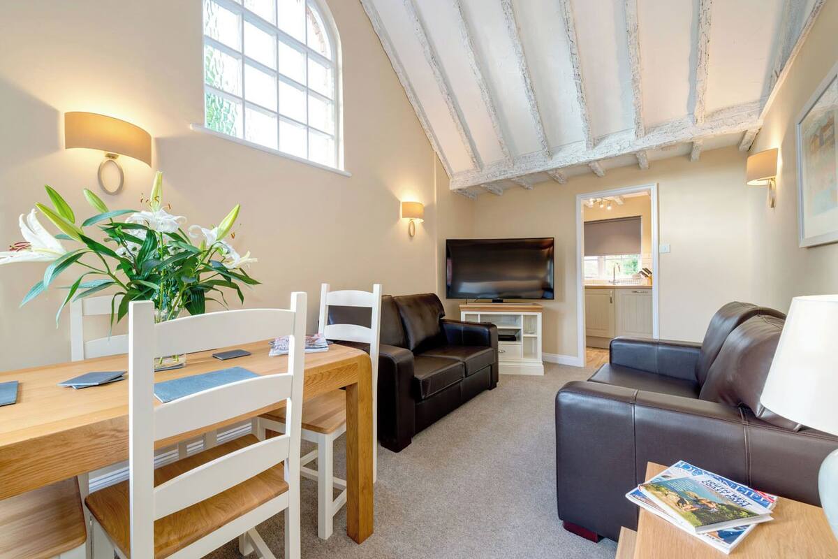 Ein gemütliches Wohnzimmer mit einem Holzesstisch, zwei Sofas, einem Fernseher und einer Küchenzeile im Hintergrund.