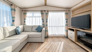 Una sala moderna con un sofá seccional, televisor de pantalla plana, piso de madera y ventanas amplias con cortinas.