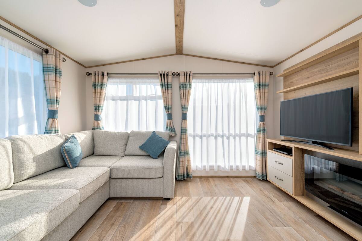 Ein modernes Wohnzimmer mit einem Ecksofa, einem Flachbildfernseher, Holzböden und großen Fenstern mit Vorhängen.