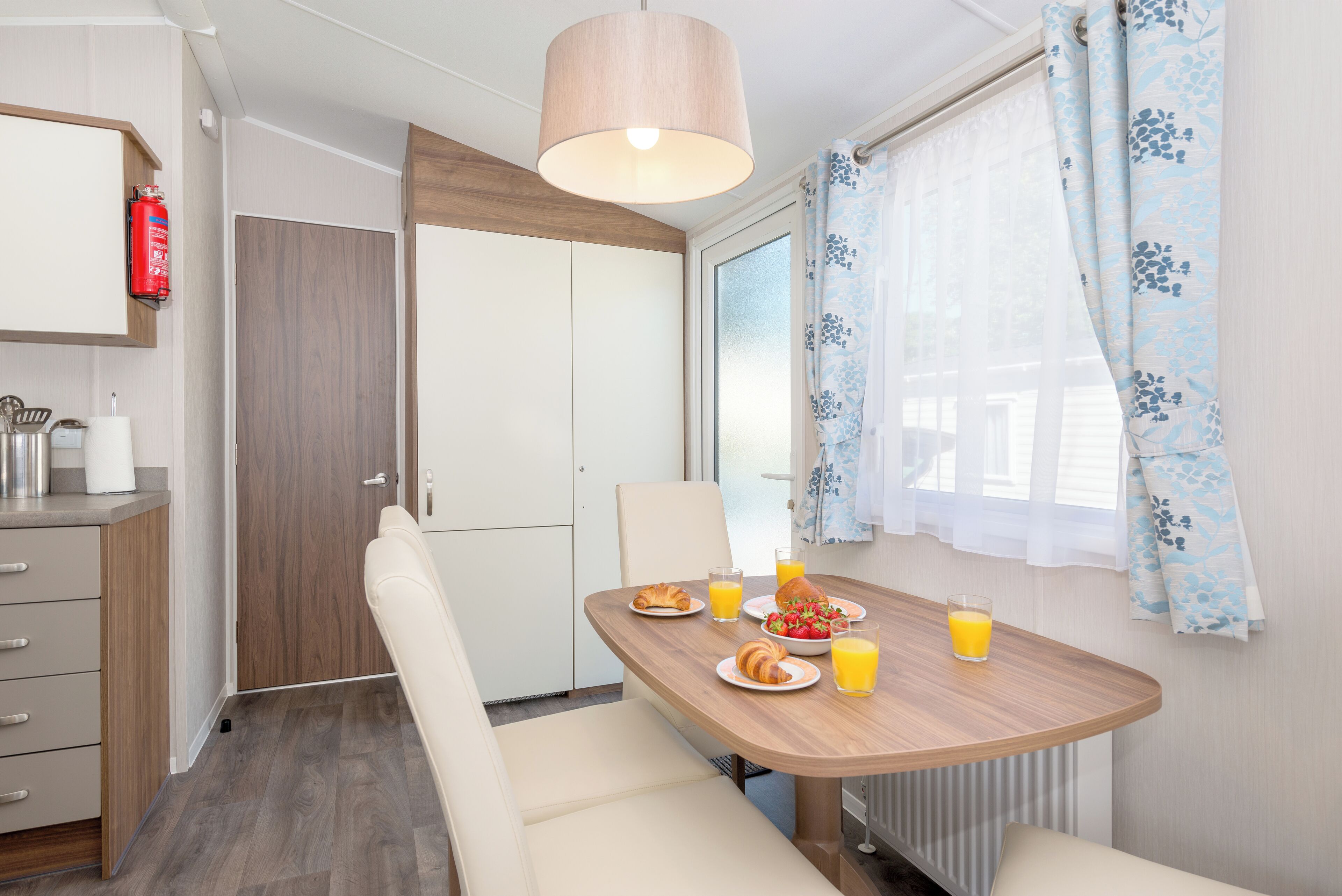 Una mesa de desayuno con cruasanes, un bol de fruta y zumo de naranja en una habitación con techo de madera, una silla blanca y una ventana con cortinas azules.
