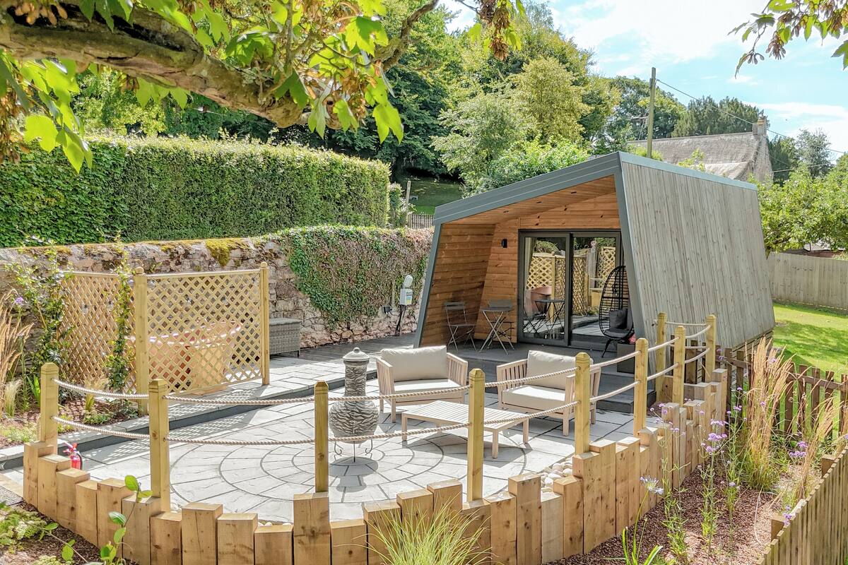 Eine Holzterrasse mit Gartenmöbeln, ein kleines Holzgebäude und eine Steinsäule.