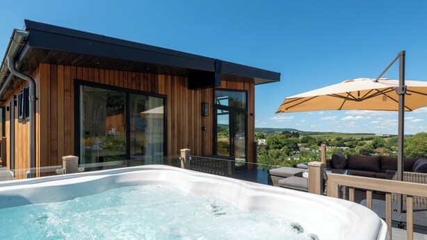 Un área moderna de hidromasaje al aire libre con cubierta de madera, un salón con cerramientos de vidrio y vistas al paisaje.