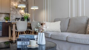 Uma sala de estar moderna com um sofá, uma mesa de centro com livros e um vaso, e uma cozinha ao fundo.