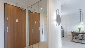 Quarto de hotel moderno com porta de madeira, divisória de vidro, área de jantar com mesa e cadeiras, e luminária pendente.