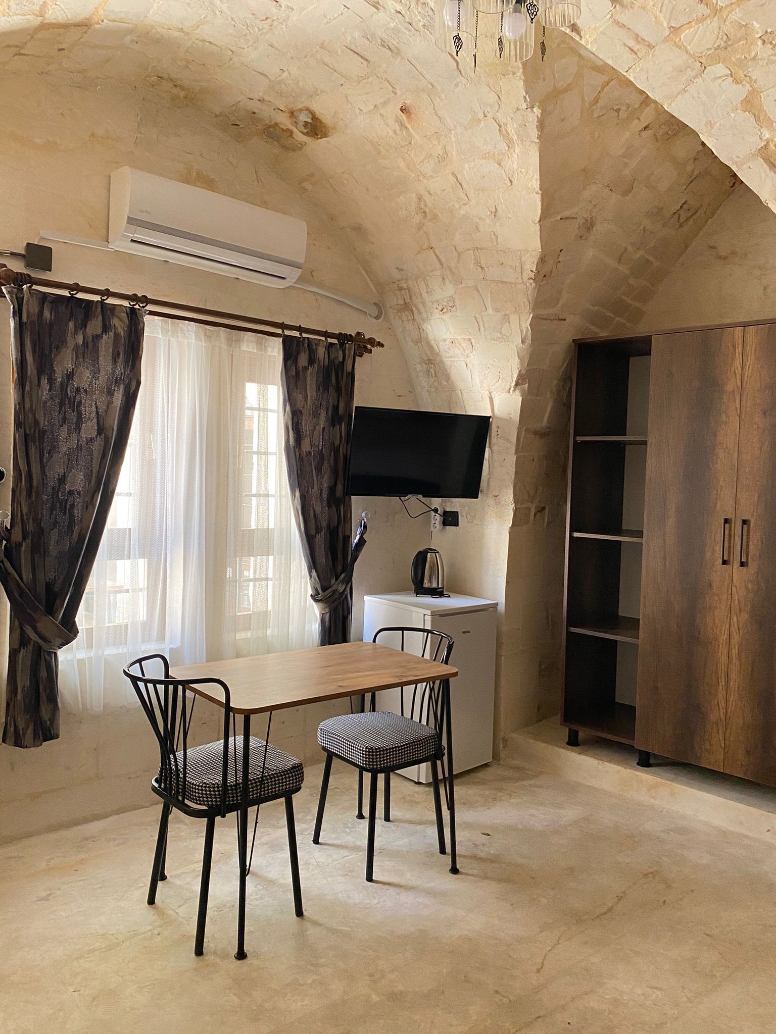 Ein Zimmer mit Steindecke, einem Holztisch mit zwei Stühlen, einem weißen Minikühlschrank, einem an der Wand montierten Fernseher und einem Holzschrank.
