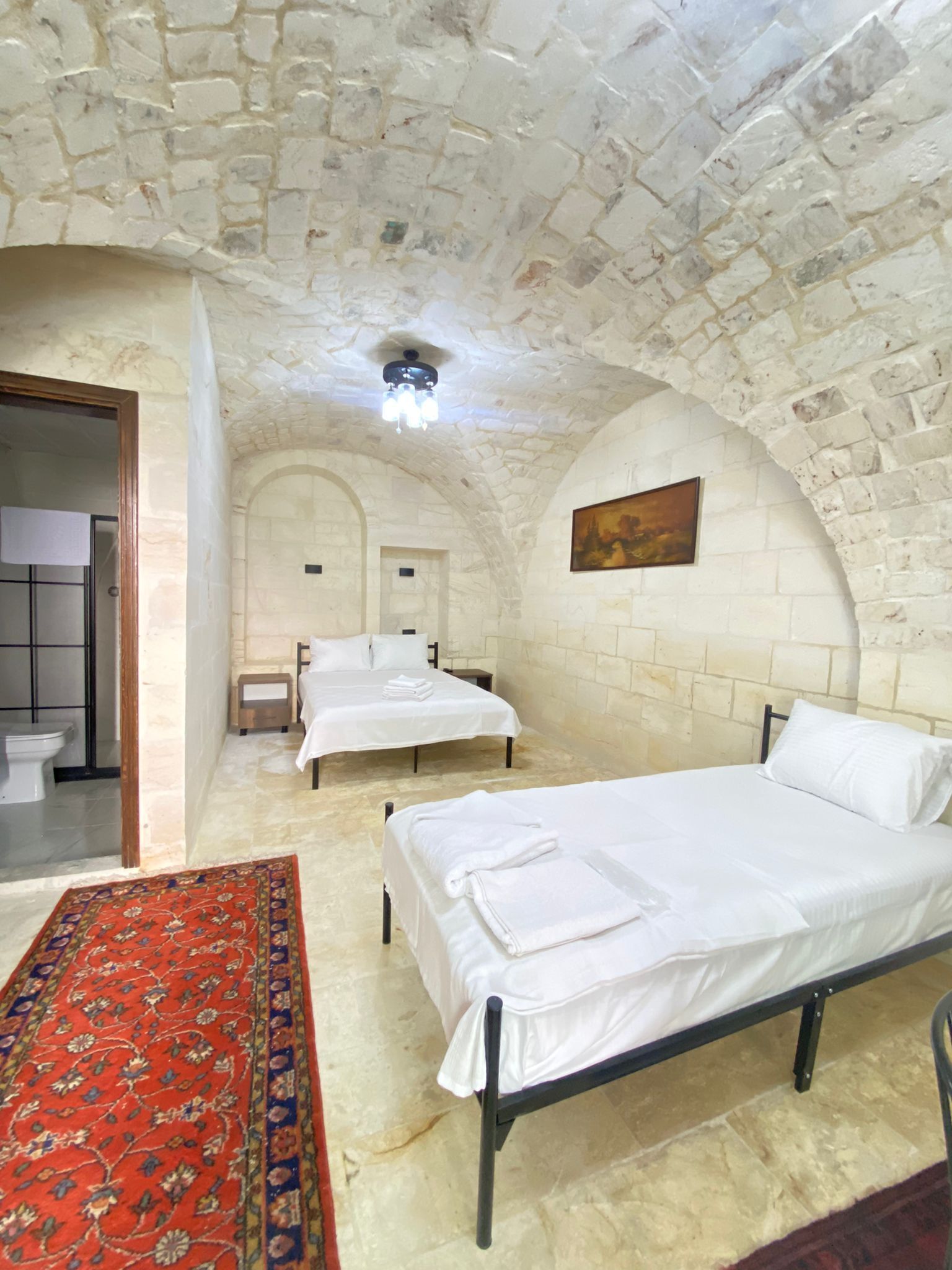 A room with two beds, a red patterned rug, stone walls, and a ceiling light.