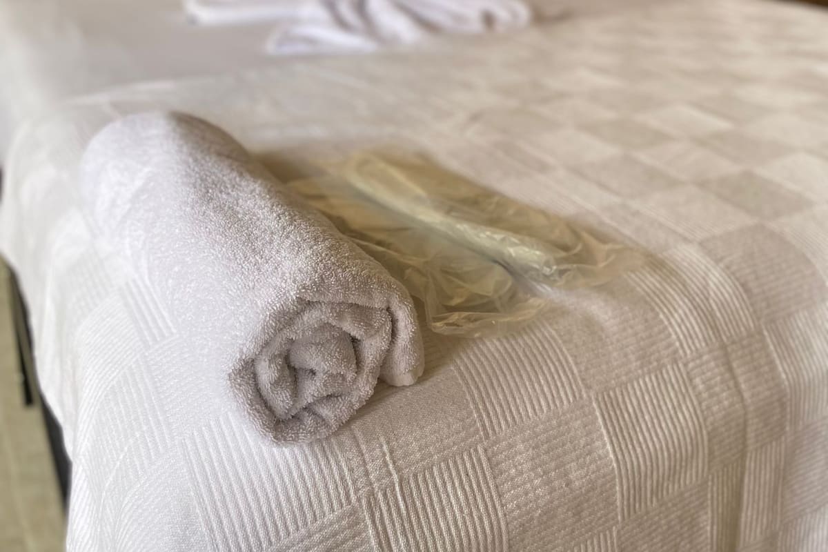 A neatly made bed with a rolled towel and a folded blanket in a room with a stone wall and wooden shelving.
