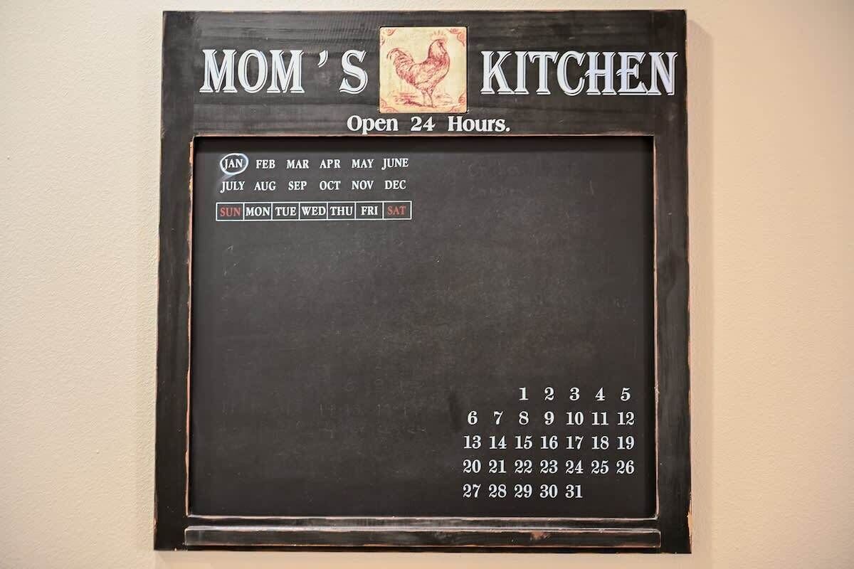 Een kalender aan de muur, met de tekst 'MOM'S KITCHEN' en 'Open 24 Uren', voorzien van een haan icoon en een maandelijks overzicht met dagen van de week.