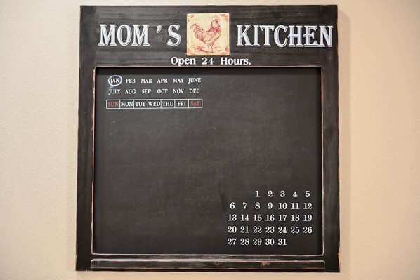 Calendario colgante con el texto 'COCINA DE MAMÁ' y 'Abierto las 24 horas', que presenta un icono de gallo y un diseño mensual con los días de la semana.