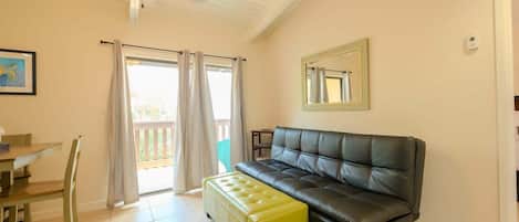 Un salon avec un canapé en cuir noir, un pouf jaune, une table à manger avec des chaises et un ventilateur de plafond.