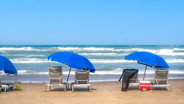 Une plage avec trois chaises de plage en métal, placées sous des parasols bleus, une glacière rouge et un sac noir sur le sable.
