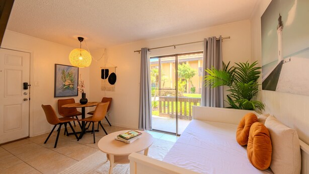 A hotel room with a bed, a dining table with chairs, a small round table with a book, a hanging light fixture, and a view of the outside balcony. - Flamingos Hideaway  (South Padre Island)