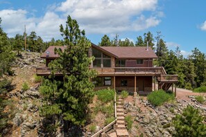 A two-story log cabin with a balcony, surrounded by trees and rocky terrain.