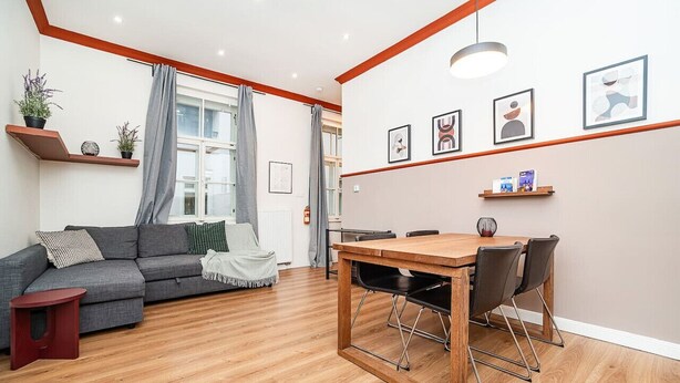 Una sala de estar moderna con un sofá gris, una mesa de comedor de madera con cuatro sillas y un taburete rojo pequeño. Hay plantas en macetas sobre una estantería, una ventana con cortinas y cuadros en la pared.