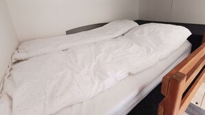 Ein Einzelbett mit weißer Matratze und weißer Bettdecke, ein Holz-Kopfteil und eine schlichte weiße Wand.