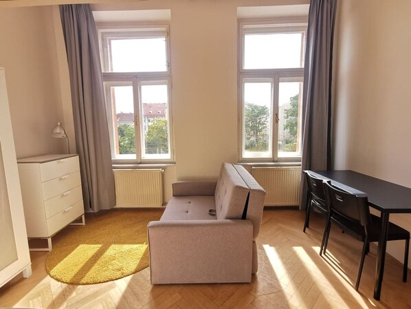 Une pièce comprenant un canapé, un bureau et une commode. On y trouve des fenêtres avec des rideaux, et un radiateur est visible.