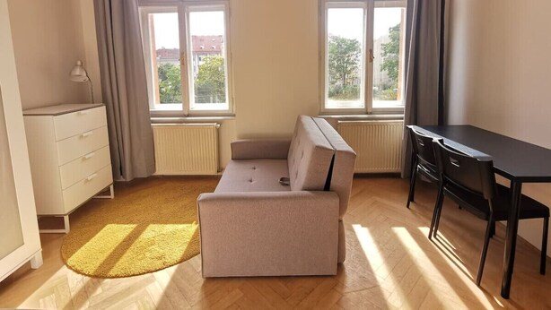 Ein Zimmer mit einer Couch, einem Schreibtisch und einem Schminktisch. Es gibt Fenster mit Vorhängen, und ein Heizkörper ist sichtbar.