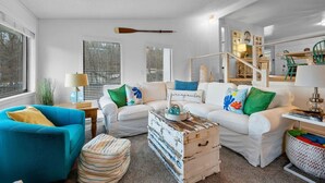 A living room with white sofas, a blue armchair, a wooden chest, and a staircase leading to a loft area. - Anchors Away-Private Dock & Slip + Fire Pit + Kayak + Grill (Sunrise Beach)