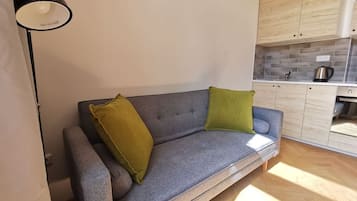 A modern living room with a grey sofa, two cushions, a black floor lamp, and a kitchen area with wooden cabinets and a tiled backsplash.