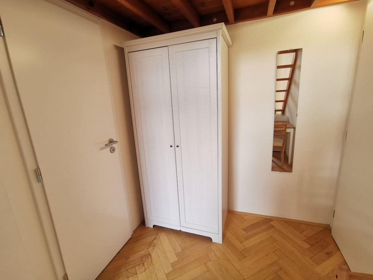 Habitación de esquina con un armario blanco, suelo de madera y una pequeña ventana con una escalera de madera.