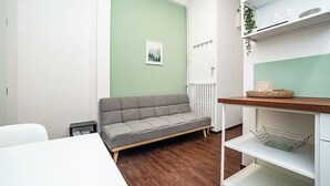 Ein Zimmer mit grauem Sofa, weißem Tisch, weißem Heizkörper und einem Regal mit Korb und Pflanze.