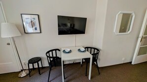 Pieni huone, jossa on valkoinen pöytä, kaksi mustaa tuolia, seinälle asennettu televisio, kehystetty kuva ja valkoisella kehyksellä varustettu ikkuna.