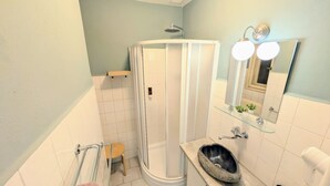 Un baño con cabina de ducha de vidrio, un lavabo con lavamanos de piedra, un taburete pequeño y un espejo con iluminación.